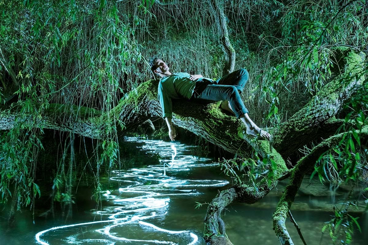 Sam Lee lies on a tree branch over an illumiated river in the dark 
