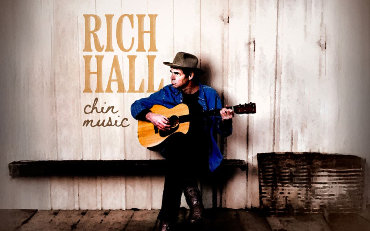 A man sits on a bench in front of a wooden wall playing guitar wearing a hat and blue denim shirt and looking off to his right. The words Rich Hall chin music appear on the left hand side of the image
