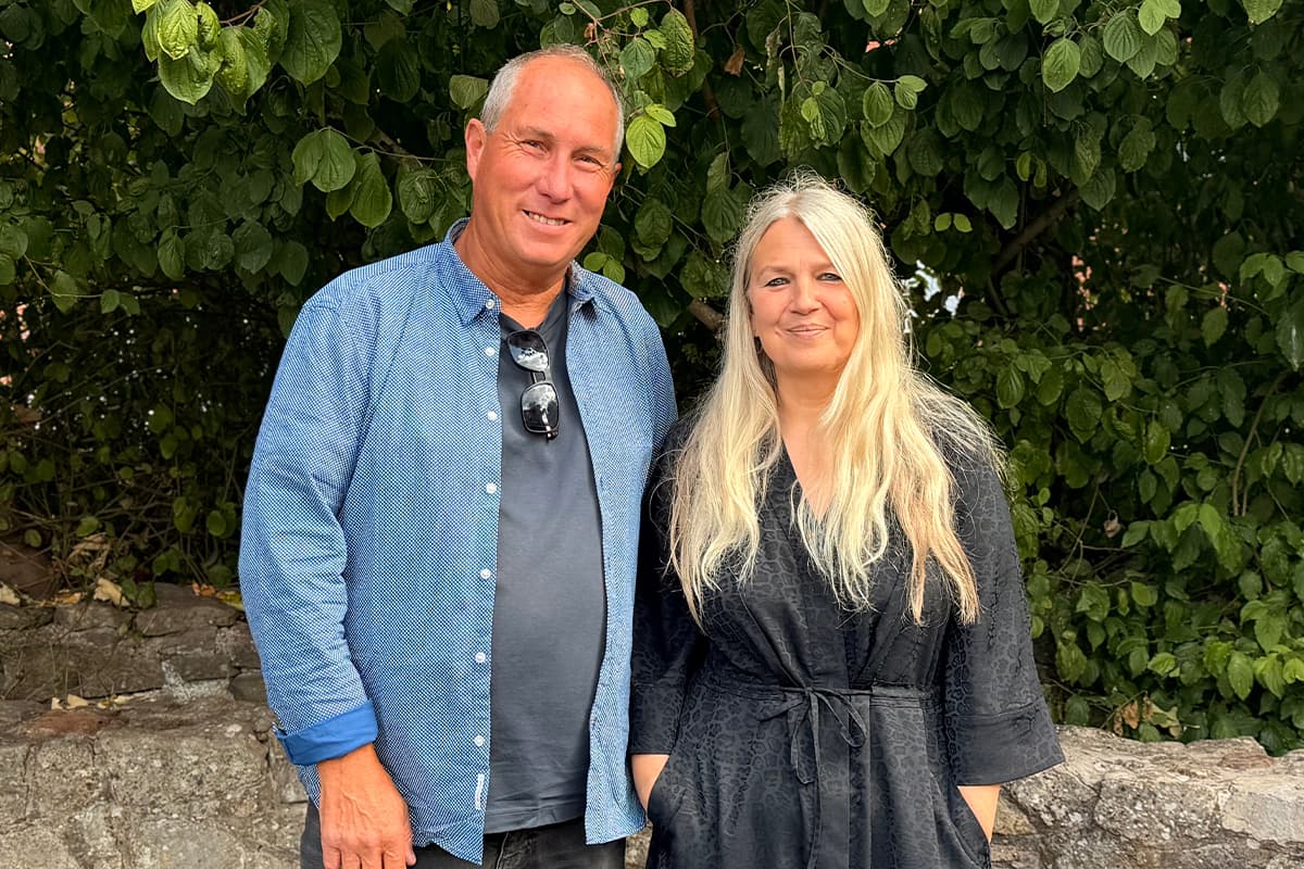 A man in a blue shirt and a blonde woman in a black dress stand smiling togther in front of a leafy backdrop